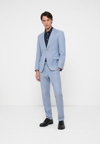 Šviesiai mėlynas kostiumas su pritaikytu siluetu, vieno eilių sagų švarkas, tamsi vienspalvė marškiniai po juo ir juodi elegantiški batai.