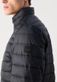 Man wearing a black quilted jacket with a raised collar and a BOSS logo patch on the upper sleeve against a plain background.