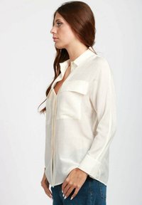 Camicia button-up di colore crema realizzata in tessuto leggero, con due tasche frontali e una vestibilità rilassata, maniche lunghe abbinate ai polsini.