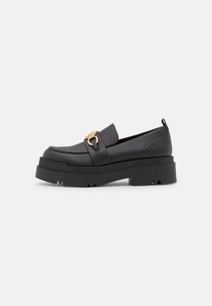 Mocassin à plateforme en cuir noir avec semelle épaisse, détail boucle dorée et nom de la marque embossé autour du bord de la semelle, vue latérale.