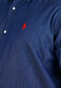 Dark blue denim shirt featuring a button-down collar, white buttons, and red embroidered logo on the chest. Smooth texture.