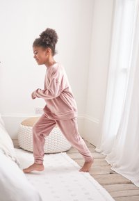 Soft pink velour loungewear set featuring a long-sleeve top and slim-fit pants. Smooth texture, relaxed design, and barefoot on wooden floor.