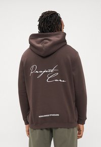 Hombre con rastas que lleva una sudadera marrón con el texto "Project Core" y "Worldwide Standard" en la parte de atrás, combinada con pantalones verdes.