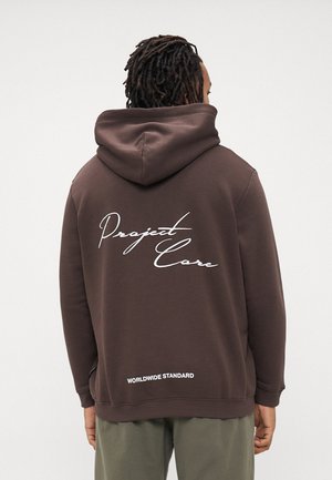 Man with dreadlocks wearing a brown hoodie with "Project Core" and "Worldwide Standard" text on the back, paired with green pants.