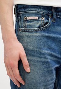 Svijetloplave traperice s blagim istrošenim finišem, koje na zadnjem džepu imaju crveno-bijeli oznaku "Calvin Klein".