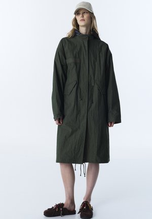 Donna in piedi che indossa un lungo cappotto verde scuro, un berretto beige e scarpe slip-on marroni con perline colorate su sfondo bianco.