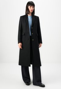 Donna con capelli scuri e lisci indossa un lungo cappotto nero sopra un maglione azzurro chiaro, pantaloni ampi neri e mocassini neri, in piedi contro un muro bianco.