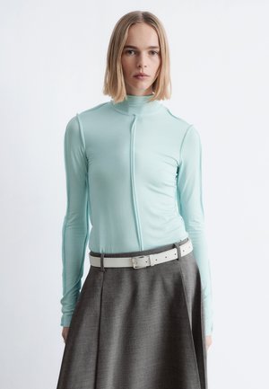 Kvinde med glat blondt hår iført en lyseblå, langærmet rullekravestrik og højtsiddende grå nederdel med hvidt bælte, stående mod en hvid baggrund.