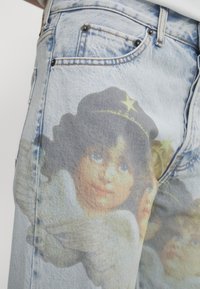 Jeans in denim chiaro con volti angelici cherubini stampati e corone d'oro a forma di stella nella zona della coscia e dell'anca.