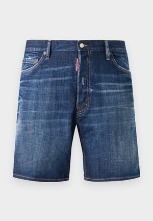 Muž stojaci pri jednofarebnej stene, oblečený v červenom tričku DSQUARED2 Milano, modrých rifľových kraťasoch, béžových ponožkách a bielych teniskách so zlatými pruhmi.