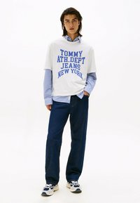 Hvid t-shirt med blå tekst, der læser "TOMMY ATH.DEPT JEANS NEW YORK" over en ternet blå skjorte, parret med mørke jeans og sneakers.