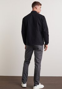 Giacca nera leggera con un design semplice, abbinata a pantaloni grigi slim-fit e sneakers bianche, vista da dietro.