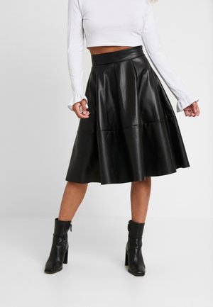 Vrouw met een zwarte leren midi-rok, een witte crop top met lange mouwen en zwarte enkelhoge laarzen met hakken, staande op een witte vloer.