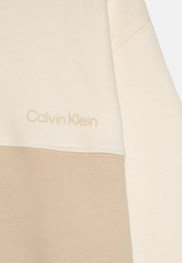 Lys beige hettegenser med teksturert overflate, med et kremfarget toppanel. Diskret "Calvin Klein" logo preget i matchende farge.