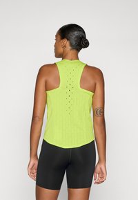 Top senza maniche verde neon con un design testurizzato, caratterizzato da perforazioni verticali e un orlo curvo. Sotto si indossano pantaloni ciclismo neri.