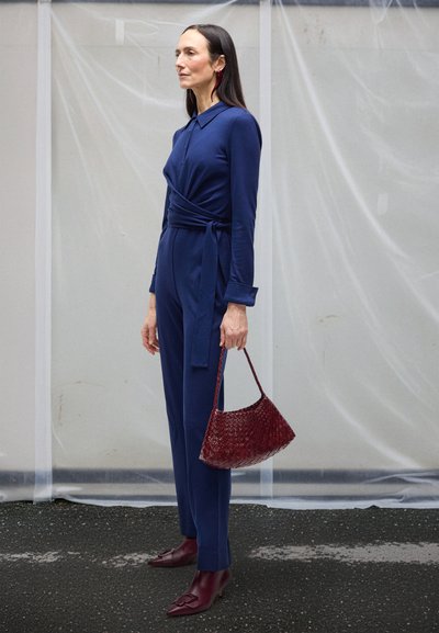 Vrouw staand in een marineblauwe jumpsuit met ceintuur, met een rode geweven handtas, rode enkellaarzen die erbij passen en lange rode oorbellen.