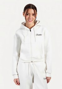 Sweat à capuche blanc court zippé, avec une capuche doublée, deux poches avant et un détail logo noir sur la poitrine. Matière douce avec une texture lisse.