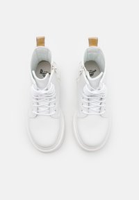 Un par de botas Dr. Martens de cuero blanco con cordones blancos y tiradores beige, vistas desde arriba sobre un fondo liso.