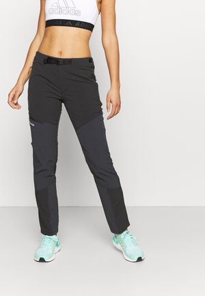 Vrouw die zwarte outdoorbroek met ingebouwde riem draagt, een wit Adidas crop top en teal sportieve schoenen, staand tegen een witte achtergrond.