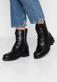 Stivali alla caviglia in pelle nera con lacci e zip. Suola robusta, tacco piatto e texture liscia. Presenta dettagli in metallo argentato.