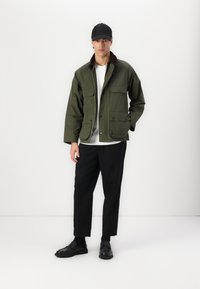 Olive green canvas jacket with brown collar, two front pockets, zipper closure, worn over a light gray shirt and black trousers with loafers.