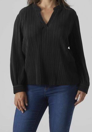 Frau trägt eine schwarze, langärmelige, plisseeartige Bluse mit V-Ausschnitt und blaue Jeans, steht vor einem schlichten grauen Hintergrund.