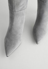 Botas de tobillo de ante gris con punta afilada, que presentan una textura suave y un diseño elegante. Detalle de cremallera en el costado.