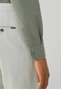 Chemise à manches longues en tissu olive texturé, dotée de poignets à boutons et d'une étiquette logo visible sur un pantalon gris clair.