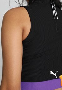 Czarny prążkowany crop top z wysokim kołnierzem, zamkiem błyskawicznym z tyłu oraz białym logo Pumy w dolnym lewym rogu. Dolny brzeg jest fioletowy.