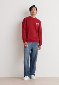 Sweatshirt rosso con colletto girocollo e grafica bianca sul petto, abbinato a jeans azzurri chiari e sneakers bianche. Sfondo semplice.