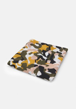 Carré de tissu avec un motif camouflage dans des tons jaunes, verts, noirs, blancs et pêche, plié sur un fond blanc.