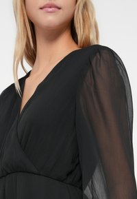 Eine Frau trägt ein schwarzes Wickelkleid mit transparenten langen Ärmeln und einem V-Ausschnitt, der ihren Hals und einen Teil ihrer blonden Haare zeigt.