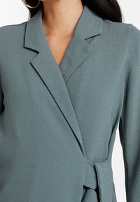 Světlý tyrkysový blejzr s texturovaným povrchem, s klopovým límcem a zavazováním v pase, představující čisté linie a přiléhavý střih.