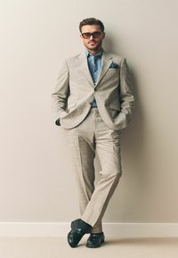 Traje a cuadros gris claro, camisa azul debajo, zapatos negros y gafas de sol oscuras. El pañuelo en el bolsillo añade detalle. Un hombre se apoya contra una pared.