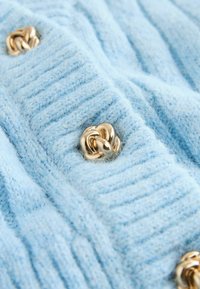 Gaiši zils adīts audums ar vertikālu ribotu tekstūru, kurā izceļas zeltaini izsukti mezgli kā ievērojami akcenti.