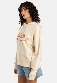 Felpa beige con un fit rilassato, caratterizzata da un ricamo di un tramonto e una palma in colore ruggine. Maniche lunghe e tasca a marsupio.