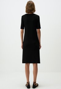 Abito nero fino al ginocchio con maniche corte e scollatura tonda. Texture liscia, forma aderente e design minimalista. Indossato con sandali neri.