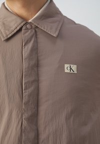Chemise marron à boutons avec une finition texturée, dotée d'un grand col et d'un petit patch logo cK sur la poitrine gauche.