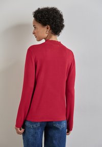 Rotes Longsleeve-Oberteil mit geripptem Ausschnitt, glatter Textur und lässiger Passform. Kombiniert mit hochgeschnittenen blauen Jeans mit kleinen Taschen auf der Rückseite.