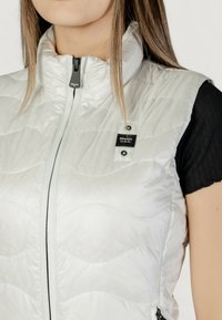 Bílá nadýchaná vesta s vysokým límcem, prošívaným vzorem, předním zipem a černou akcentní nášivkou. Nošena přes černý top s krátkým rukávem.