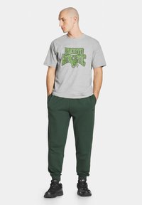 Szara bawełniana koszulka z zielonym napisem "PROSTO ATHLETIC", w połączeniu z ciemnozielonymi spodniami dresowymi i czarnymi butami sportowymi o teksturowanej powierzchni.