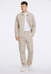 Beige bomullsskjorta med två bröstfickor och en vit T-shirt under, i kombination med matchande beiga byxor och vita sneakers.