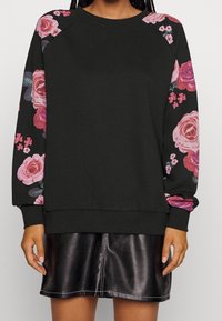 Černá mikina s květinovými vzory v růžové a fialové na rukávech; s kulatým výstřihem a žebrovanými manžetami. Nošená přes černou koženou sukní.