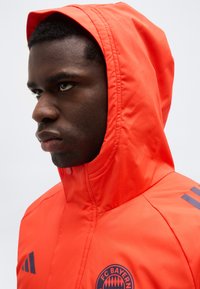 Mladi moški v svetlo oranžni kapuci FC Bayerna, obrnjen levo z osredotočenim izrazom na obrazu, pred enobarvnim ozadjem.