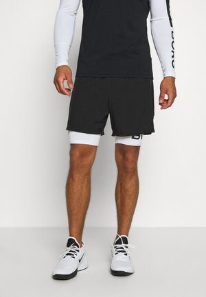 Sportowiec ubrany w czarne spodenki na białych krótkich spodenkach kompresyjnych, czarno-biały sportowy top z długim rękawem oraz białe buty sportowe z czarnymi akcentami.