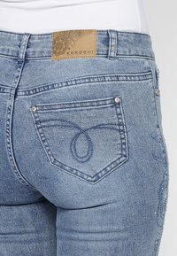 Vue de dos d'un jean en denim bleu clair avec un motif de poche brodé et un patch de marque en cuir marron à la taille.