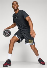 Camiseta negra de manga corta con un logo blanco, pantalones cortos negros con ribetes blancos y el número "24" en amarillo, sosteniendo un balón de baloncesto negro texturizado.