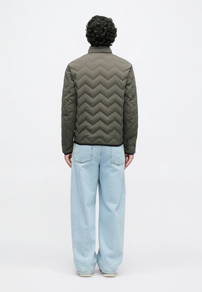 Olivovo zelená prešívaná bunda so vzorom šípiek, s vysokým golierom a čiernym lemovaním na spodku, v kombinácii so svetlomodrými džínsami s širokými nohavicami.