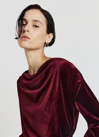 Bordeauxrotes Samtoberteil mit drapiertem Ausschnitt, langen Ärmeln und einem sanften Glanz. Verfügt über eine entspannte Passform und eine glatte Textur.