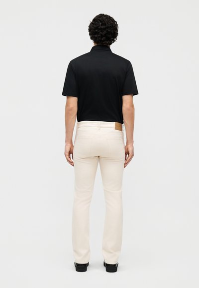 Άνδρας με σγουρά μαλλιά στέκεται πλάτη, φορώντας μαύρο πόλο μπλουζάκι με κοντά μανίκια, εκρού τζιν και μαύρα παπούτσια σε απλό φόντο.